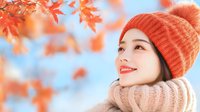 Young Asian woman wearing orange knit beanie and cozy turtleneck sweater smiling while looking up at falling autumn maple leaves against blue sky background.