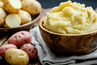 Creamy mashed potatoes served in wooden bowl with fresh raw red and yellow potatoes on dark background, rustic food styling for restaurant menu.