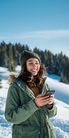 Young woman in green winter parka and knit beanie smiling while using smartphone against snowy mountain landscape with pine forest background.