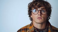 Young Caucasian man with curly brown hair and round glasses looking up with wonder expression, wearing plaid yellow flannel shirt against light blue background.