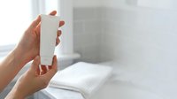 Close-up of female hands holding blank white cosmetic bottle in modern bathroom. Product mockup for skincare, lotion, or shampoo packaging display.