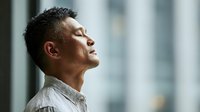 Asian man with closed eyes in profile against blurred office background, expressing moment of calm reflection or meditation during workday.