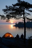 Tranquil lakeside camping scene with illuminated tent and two people silhouetted against sunset sky, pine tree framing serene water view and rocky shoreline.