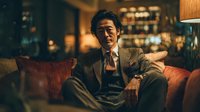 Confident middle-aged Asian businessman in elegant suit relaxing on luxury leather sofa in upscale lounge with warm ambient lighting and sophisticated interior.
