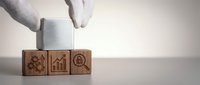Hand placing wooden cube with gear icon completing business strategy concept with growth chart and target symbols on neutral background.