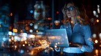 Professional businesswoman working late on laptop in modern office with city lights bokeh background for corporate presentations and business concepts.