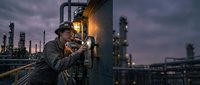 Industrial worker in safety gear operating equipment at oil refinery during twilight with dramatic sky and industrial complex background.