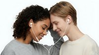 Young multiracial couple sharing earphones, heads touching, smiling with eyes closed, enjoying music together in casual outfits on white background.