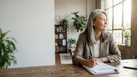 Mature woman with gray hair and Caucasian ethnicity, wearing a beige blazer and white shirt, sits at a wooden desk in a bright, plant-filled office space, writing on a notepad while gazing thoughtfully toward a window, suggesting professional contemplation and business planning.