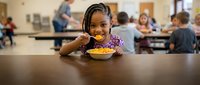 Young African American girl with braided hair enjoying healthy school lunch meal in busy cafeteria with students and staff in background.