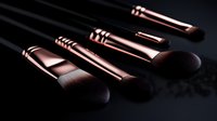Professional makeup brushes with rose gold metal handles and soft brown bristles arranged on dark background with dramatic lighting and reflective surface.