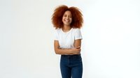 Young African American woman with curly afro hair smiling confidently in white t-shirt and jeans against clean background for lifestyle marketing.