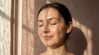 Young woman with East Asian ethnicity, eyes closed, serene expression, standing by window with natural sunlight casting shadows on neutral beige wall, skincare and wellness concept.