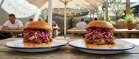 Two gourmet pulled pork sandwiches with purple cabbage slaw on brioche buns served on white plates at outdoor restaurant patio dining setting.