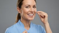 Young woman with light skin tone and brown hair wearing light blue scrubs, smiling while holding a dental mirror near her mouth against a gray background. Suitable for dental, healthcare, medical professional, and dentistry-related content.