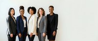 Diverse group of five professional women standing together against white background, wearing business suits and blazers, representing corporate teamwork, leadership, and workplace diversity for business and corporate communications.