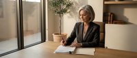 Professional mature Caucasian businesswoman in dark blazer reviewing documents at modern office desk near large windows with natural lighting and plants.