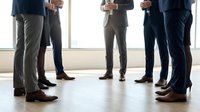 Corporate professionals in business attire standing in circle formation during meeting or team building event in modern office space with large windows and natural light.