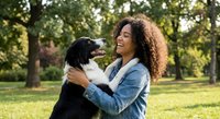 Happy young African American woman with curly hair embracing black dog in sunny park. Joyful pet owner bonding with loyal companion outdoors on beautiful day.