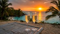 Two champagne glasses on wooden deck overlooking tropical beach sunset with palm trees and turquoise water creating romantic vacation atmosphere.