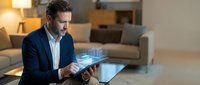 Caucasian businessman in a suit using a tablet with a glowing holographic city projection in a modern living room interior.