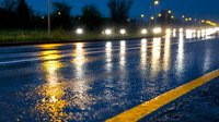 Wet asphalt road at night with glowing street lights reflecting on rain-soaked surface. Yellow line marking visible. Atmospheric urban scene for transportation themes.