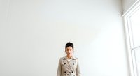 Young Asian woman wearing a beige trench coat standing in a bright, minimalist white interior space with natural light from window, suitable for corporate and lifestyle design projects.