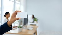 Close-up of male hand holding blank white business card in modern office workspace with colleagues working at desk in background.