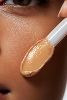 Makeup artist applying liquid foundation with applicator on African American woman's face, showing texture of cosmetic product and glowing skin close-up.