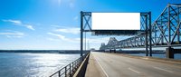 Steel truss bridge spanning wide river with concrete roadway and metal railings under bright blue sky with white clouds on sunny day.