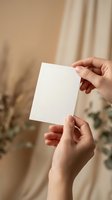 Female hands holding blank white paper card mockup in warm natural light with soft beige curtain background for branding and stationery design.