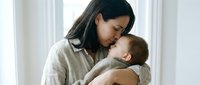 Asian mother tenderly holding newborn baby in soft natural light, intimate bonding moment for parenting and family lifestyle content.