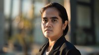 Young Asian businesswoman with determined expression in black blazer against blurred urban background during golden hour lighting.