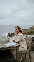 Middle-aged woman working remotely on laptop at wooden table overlooking rocky coastline and ocean. Remote work, freelance professional, coastal lifestyle.