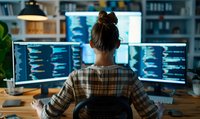 Female software developer in plaid shirt working late at multiple computer screens showing code, view from behind in dark office with ambient blue monitor glow.