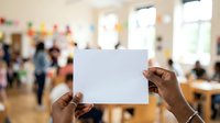 Close-up of hands holding blank white card in front of blurred classroom with diverse students and teacher, suitable for educational messaging, announcements, and promotional content.