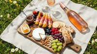 Overhead view of charcuterie board with cured meats, cheese, grapes, nuts and bread arranged on wooden serving plank on white linen cloth over green grass with wine bottle and glasses.
