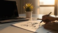 Hand drawing bicycle sketches on paper at modern workspace with laptop and plant in warm sunlight for creative design projects.