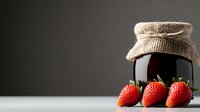 Fresh ripe strawberries next to dark glass jar with burlap lid on gray background with copy space, minimalist food composition for preserves and jam.