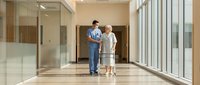 Male healthcare worker assisting elderly Caucasian woman with walker in modern medical facility corridor with natural lighting and glass walls.