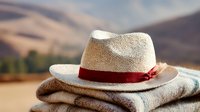 Straw fedora hat with red ribbon band resting on folded blanket against blurred mountain landscape background, capturing rustic outdoor adventure spirit.