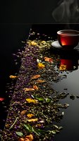 Steaming cup of black tea with scattered dried flowers, herbs and tea leaves creating artistic composition on dark reflective surface with rising steam.