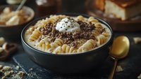 Creamy fusilli pasta topped with seasoned ground beef and sour cream in dark ceramic bowl, served on rustic surface with golden spoon, comfort food styling.