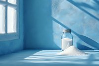 Glass jar filled with white salt and spilled pile on blue surface, dramatic window light casting geometric shadows creates minimalist composition.