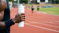 Athlete holding water bottle on running track during outdoor training session with team in background.