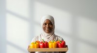 Young woman of African descent wearing white hijab holding wooden tray with fresh bell peppers in yellow and red colors against bright white background with natural window light.