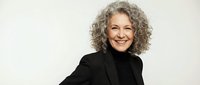 Confident mature woman with curly gray hair smiling warmly in black business attire against neutral background for professional corporate content.