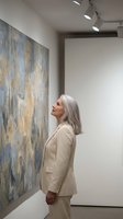Mature woman with gray hair in beige suit admiring abstract painting in modern art gallery with track lighting and white walls.