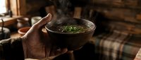 Caucasian male hand holding a steaming rustic bowl of herb broth soup in a cozy wooden cabin interior, warm moody atmosphere.