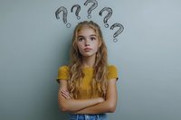 Confused young blonde woman with question marks above head looking up thoughtfully with crossed arms against gray background expressing doubt and uncertainty.